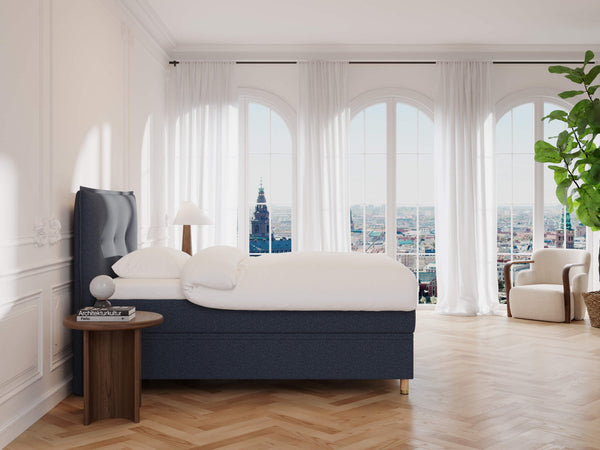 Et stilfuldt soveværelse har Alva Fjäll mørkeblå polstret sengeramme (120x200) med hvidt sengetøj. Et sidebord af træ holder en lampe og indretning ved siden af sengen. En stor plante ved et vindue med gennemsigtige gardiner inviterer til naturligt lys, suppleret med elegant sildebensgulv.