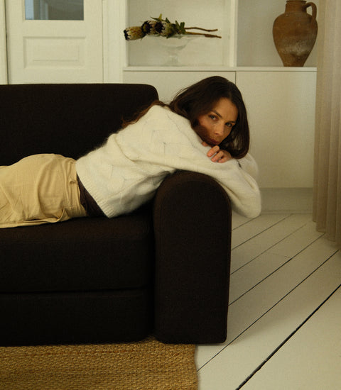 Kvinde liggende på mørkbrun sofa iført blød, hvid kabelstrikket sweater og beige bukser, neutrale interiørfarver