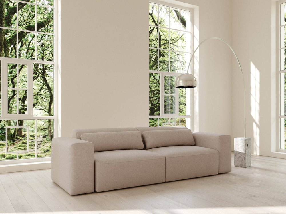 Sovesofa i macchiato-beige stof, lavt modulært design med runde armlæn og to ryghynder foran store vinduer.