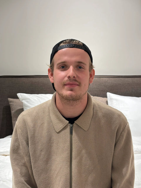 Beige strikket zip-sweater med krave på seng, hvidt og gråt hovedgærde i baggrunden, person iført kasket sidder.