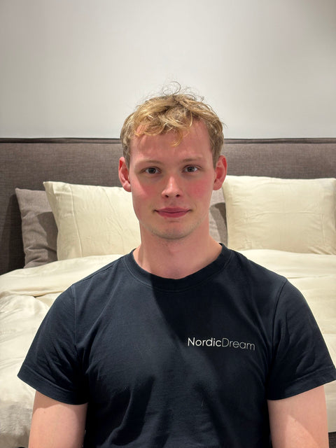 Seng med lysegrå polstret gavl og cremefarvede puder i bomuld, person iført sort NordicDream T‑shirt foran sengen