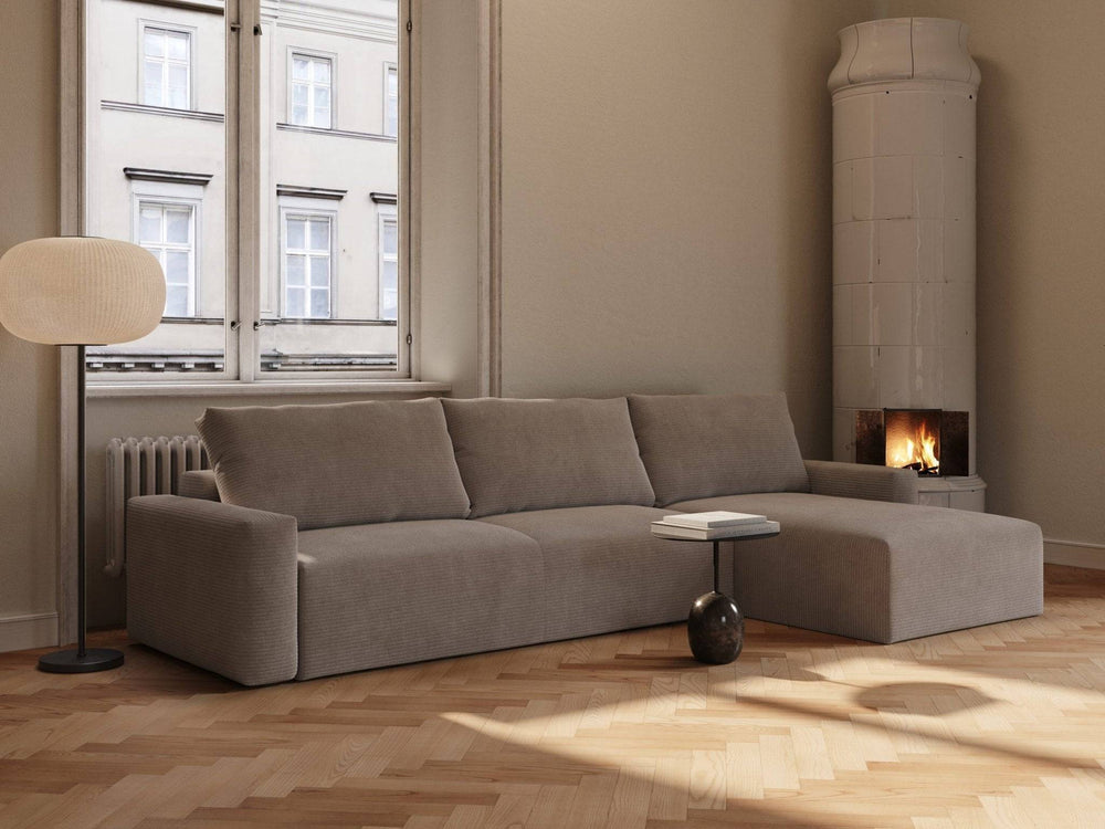 Lattefarvet fløjls sovesofa Maru 145x342 cm i L-form med bløde puder, lavt sort sidebord, parketgulv og tændt pejseovn.