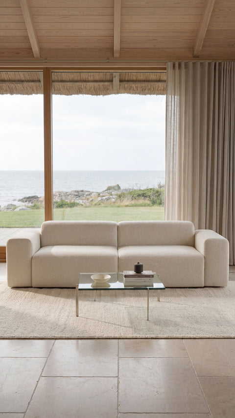 Beige modulsofa i blødt stof foran stort vindue med havudsigt, glasbord og neutrale gardiner.