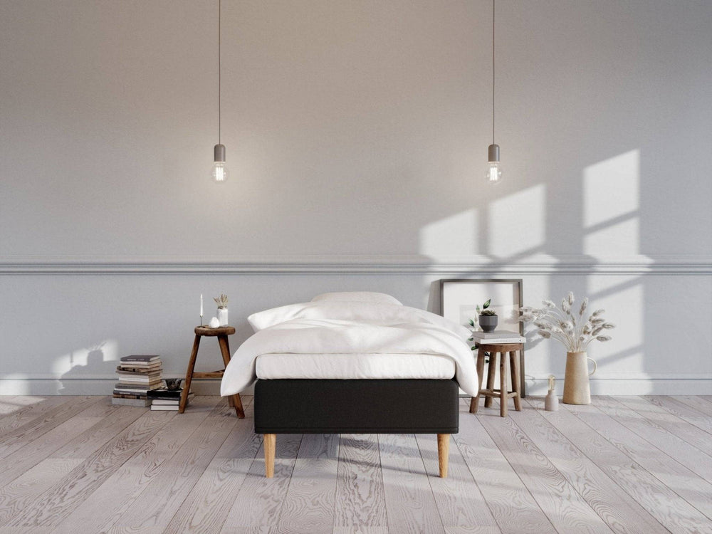 Sort boxmadras 80x200 med træben, hvidt sengetøj i lyst, minimalistisk soveværelse, dekoreret med små borde og vaser.