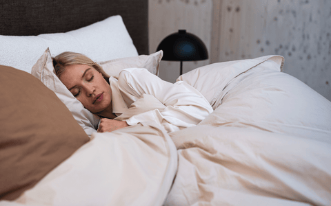 Person ligger under blødt, lysebrunt og beige sengetøj med matchende puder og mørk natbordslampe i baggrunden