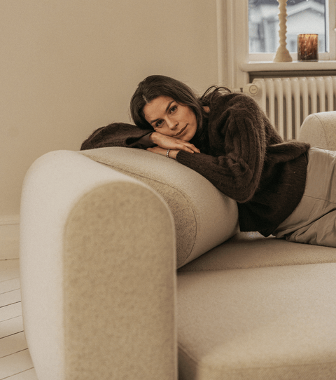 Kvinde ligger afslappet på blød beige sofa med rund armlæn iført brun uldtrøje, nær vindueskarm med dekorativt lys.