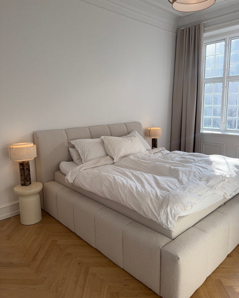 Lysegråt polstret sengehoved og boxmadras med hvidt linned, matchende natbordslamper og beige gardin ved vindue