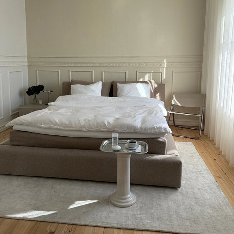 Sengen med hvidt sengetøj og store hvide puder på beige polstret sengegavl, sølvbakke med kop og glas foran