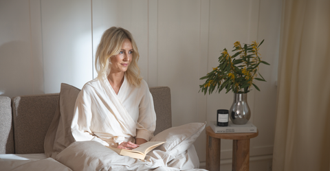 Kvinde sidder i beige bomuldsbadekåbe på lyst sengetøj, gråt sengegavl, natbord med vase og grønne grene.