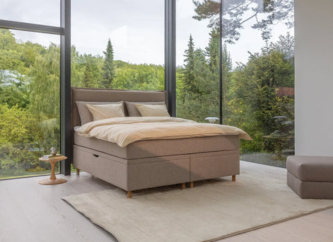 Beige polstret seng med lyse sengetøj og puder foran store vinduer med udsigt til grøn skov.