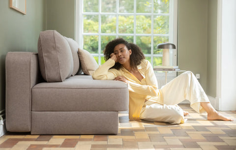 Beige-grå sofa med bløde pudebetræk i stue, kvinde læner sig mod sofaen i lys gul bluse ved stort vindue