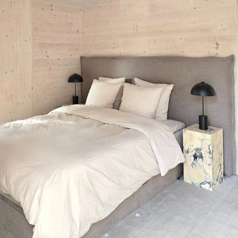 Seng med beige sengetøj og puder, gråt polstret gavl, marmornatbord og sorte sengebordslamper.