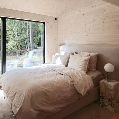Seng med beige-linned dyne og puder, polstret hovedgærde, marmorsidebord og stort vindue mod skov.