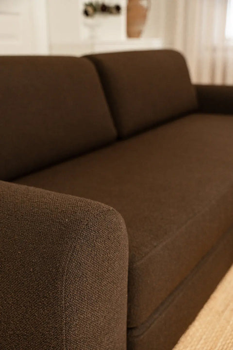 Mørkebrunt sofa-tekstil i grovvævet materiale med bløde afrundede armlæn og lige ryghynde i nærbillede