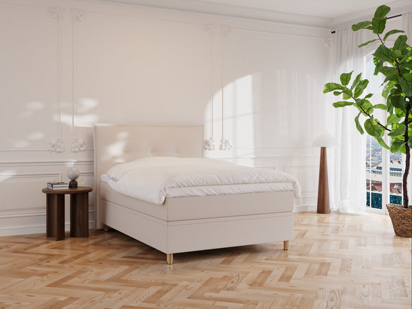 Et minimalistisk soveværelse har en Alva Fjäll-sandseng på et sildebenstrægulv. Ved siden af står et lille rundt bord med en lampe. En høj plante er tæt på vinduet med gennemsigtige gardiner, der lukker blødt naturligt lys ind.