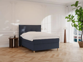 Dyb blå kontinentalseng 120x200 med tekstilpolstret hovedgærde, hvidt sengetøj, gulv af lyst træ og planter.