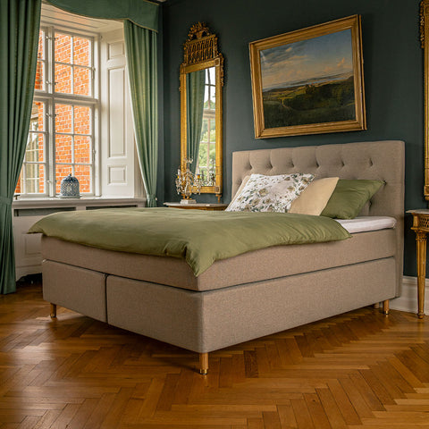 Boxmadras med beige polstret sengestel, grøn dyne og mønstret pude i elegant soveværelse.