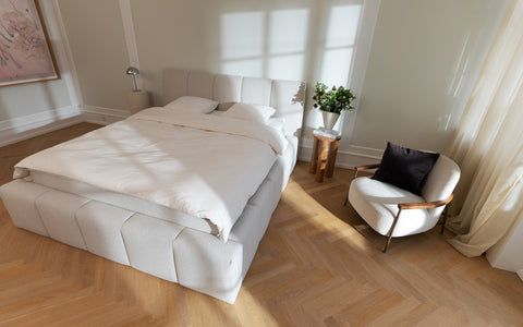 Lyst, polstret dobbeltseng i creme med blødt duvet, matchende puf, træsidebord og lyse gardiner