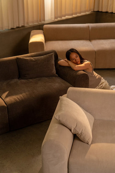 Lyse og mørkebrune sofaer i blødt, textured stof med puder; en person hviler armene på armlænet.