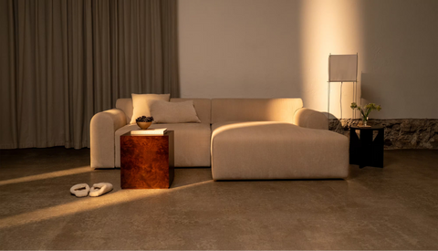 Beige stofsofa med chaiselong og pudebetræk, træsidebord, bløde hjemmesko på gulvet og varm belysning.