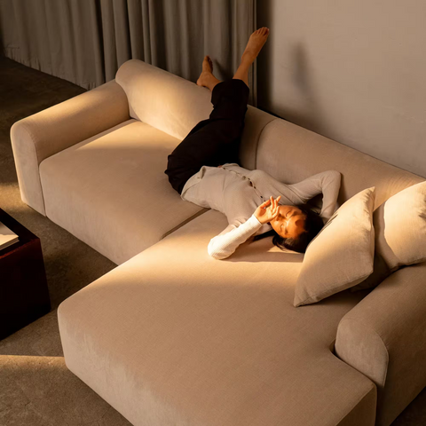 Beige stofsofa med bløde afrundede armlæn og pude, person ligger afslappet med ben op ad væggen.