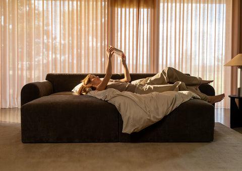 Mørkegrå sofa seng med beige sengetøj og tæppe, person ligger og læser under varmt sollys fra lysegardin.