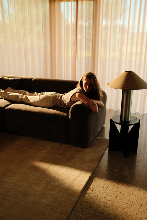 Mørkegrå sofa med beige pude og lampe, person ligger afslappet på tværs i varme, gyldne toner foran lysegardiner.