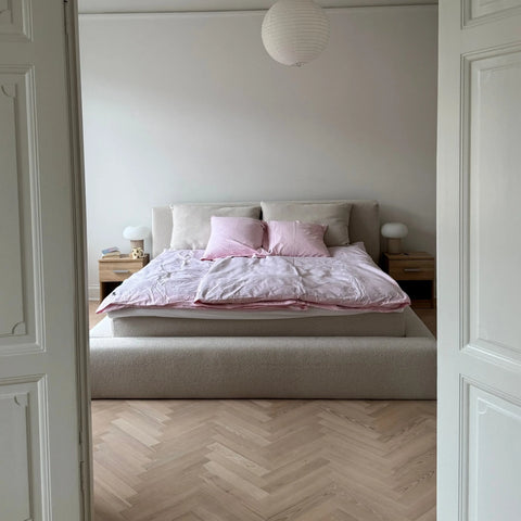 Seng med beige polstret gavl, lyserødt sengetøj og puder, to træ-natborde og papirlampe over sengen.