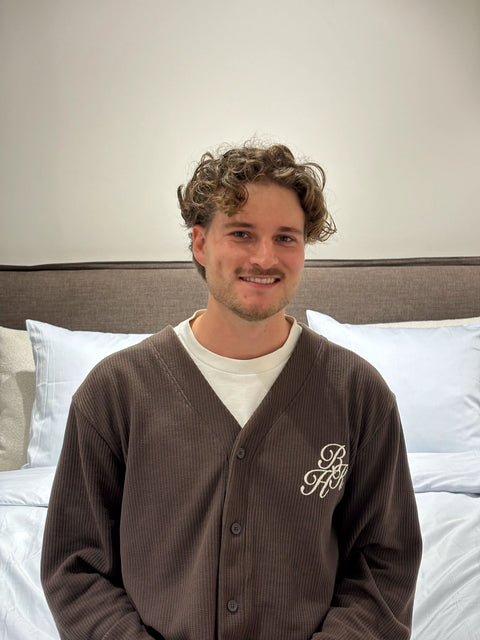 Mørkebrunt ribstrikket cardigan-lignende nattekåbe med knapper og hvit broderet monogram, person sidder på seng med lyse