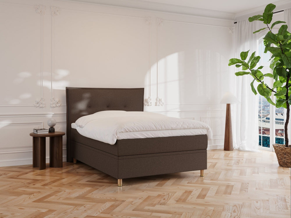 Et minimalistisk soveværelse viser Alva Nordlys sengen i mørkebrun med hvidt sengetøj. Et sidebord i træ rummer en lampe og indretning. En stor potteplante står ved vinduet, mens sollys kaster mønstre på de hvide vægge og parketgulv.