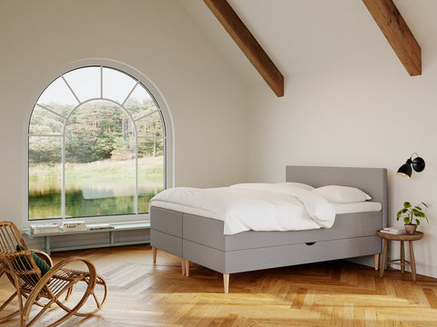 Soveværelse med grå boxmadras og hvidt sengetøj, buet vindue med sø- og skovudsigt, trægulv og rottingstol.