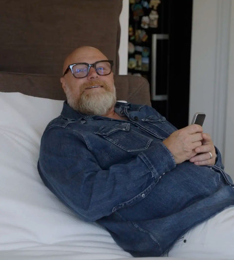 Mand i blå denimskjorte liggende i hvidt sengetøj med brun nakkestøtte, holder smartphone, nærbillede.