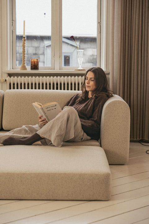 Beige boucle-sofa med afrundede armlæn, kvinde læser bog, naturfarvede gardiner og vindueskarm med stearinlys og glasvasen