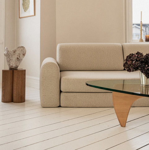 Beige tekstursofa med afrundede armlæn ved vindue, træsidebord og keramisk skulptur i neutral, varm indretning