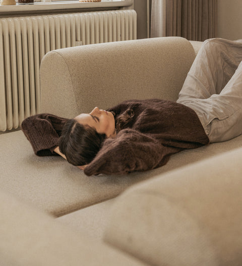 Beige sofa med blødt, vævet stof og person liggende afslappet med brun uldtrøje og lyse linnedbukser