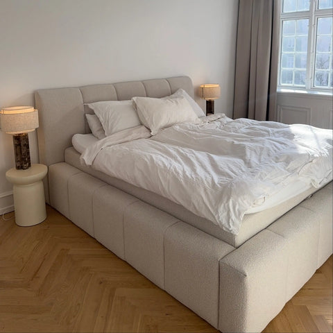 Seng med lysegrå polstret ramme og naturhvidt sengetøj, to puder og bløde dynebetræk ved vindue og bedside lamper.