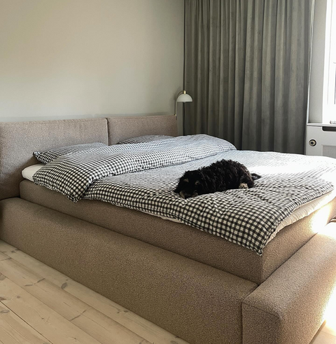 Støvet beige polstret seng med sort-hvid gingham dyne og pude, mørk hund liggende midt på sengen, grå gardin i baggrunden