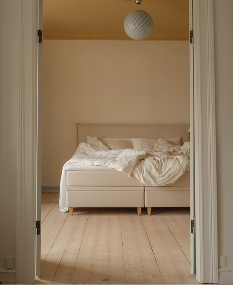 Sydd seng i beige stof med krøllede creme sengetøj, polstret hovegær og rund hvid loftslampe over.