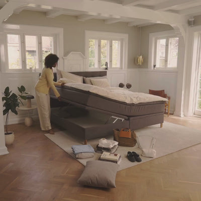 Lyst soveværelse med grå boxmadras, creme sengetøj og en person, der rejser madrassen i et lyst, nordisk rum