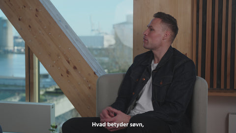 Mand sidder i grå lænestol med mørk denimjakke foran stort vindue og lyst træinteriør, havudsigt i baggrunden