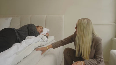 Person ligger på hvidt madrassystem med blødt, beige polstret sengegavl; konsulent viser lag og madrasstruktur.