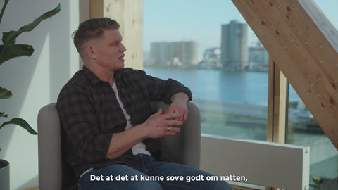 Person sidder i lysegrå stofstol ved vindue med havudsigt, træramme og grøn plante i moderne, nordisk interiør.